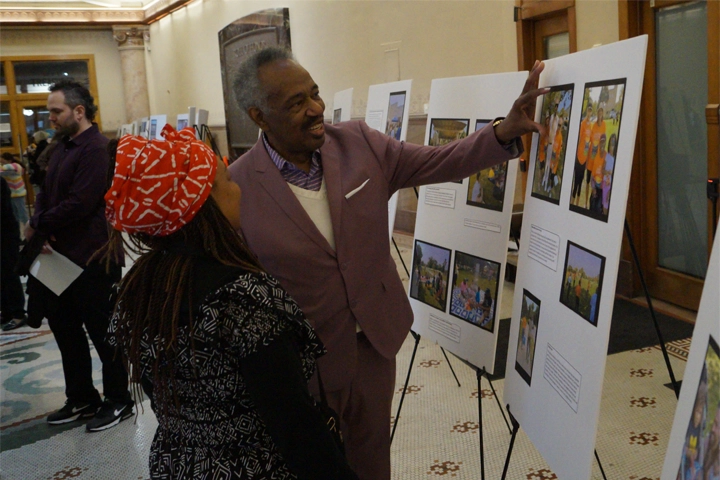 A Black man wearing a gray blazer gestures to a white board on an easel. The board is covered in photographs. A Black woman with an orange head covering looks on.
