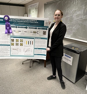 A young white woman with her hair pulled back wears a black blazer and pants. She stands next to a poster which  sports a purple ribbon.