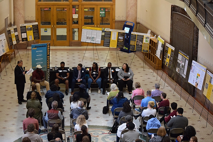 A birds-eye-view of a group of people sitting in rows of folding chairs facing a panel of people at the front of the room. Around the perimeter of the room are academic posters on easels.