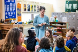 A woman with short, curly gray hair and black glasses wearing a blue dress and cardigan stands in front of seated children in a classroom.