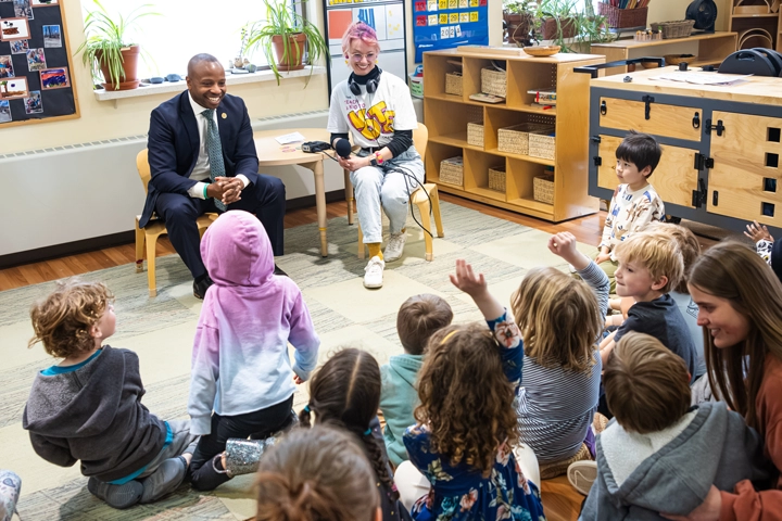 A Black man in a dark suit sits next to a younger white woman with pink hair and glasses. She wears a t-shirt that reads "Teach a Kid to Vote." They face an audience of young children sitting on the ground.