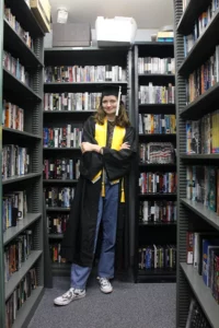 A young woman wears a black graduation gown over jeans and a t-shirt. She has a mortarboard with a tassel on her head. She stands in front of shelves of films.