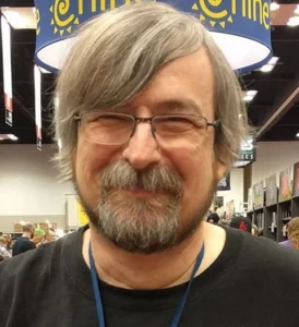 A white man with graying, shaggy hair, a graying goatee, and glasses stands in a busy convention center.