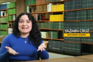 A young Hispanic woman with curly dark hair and a blue shirt gestures with her hands and looks at the camera. In the background are shelves of books.