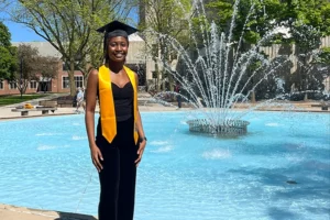 A young Black woman wearing a yellow stole and a mortarboard stands in front of a fountain and smiles at the camera.