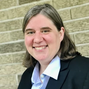 A headshot of a white woman with shoulder-length dirty blond hair. She wears a black blazer and smiles at the camera.