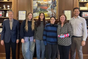 A group of people stand in a wood-paneled office. From left is an older man in a suit, three young women in casual clothes, a woman with her arm in a cast, and a man in business casual wear.