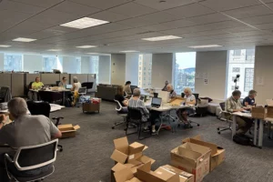 A group of people sit at tables scattered around a large office space. A stack of cardboard boxes lies in the center of the room.
