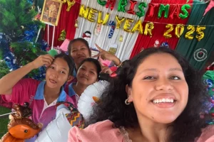 A young woman with tan skin and dark curly hair takes a smiling selfie. Three southeast Asian women smile in the background. Behind all four are balloon letters that say Merry Christmas and Happy New Year 2025.