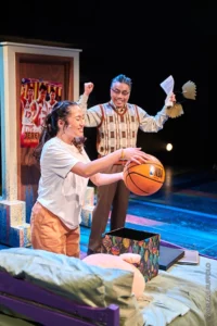 A young Asian woman holds a basketball. Behind her, an older Asian man raises his arms in triumph. The two are on stage in a bedroom set.