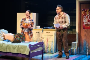 Two Asian actors stand on stage in a bedroom set. One actor is a teenage girl and the other is an older man in glasses and a sweater vest.
