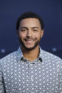 A headshot of a young Black man with a short beard. He wears a patterned button down shirt and smiles at the camera.