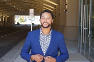 A young Black man with a short beard smiles at the camera. He wears a blue suit jacket over a patterned shirt. He is standing in front of a lit tunnel.