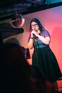 A white woman with dark hair and glasses stands on stage holding a microphone.