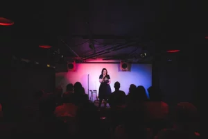 A white woman in a dress and red glasses stands on a lit stage holding a microphone as she addresses a darkened theater audience.