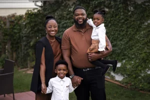 A Black woman and man stand side by side, smiling at the camera. The woman wears a brown shirt with a flowing black vest. The man wears a brown polo shirt and black pants. The couple holds the hands of a little Black boy wearing a white button-down shirt. The man holds a toddler Black girl on his hip. In the background is an ivy-covered wall.