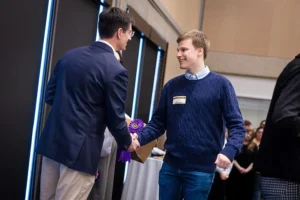A young white man with blond hair wearing a blue sweater with a nametag shakes hands with an older white man wearing a blue blazer.