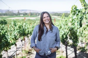 A young white woman with long brown hair and a blue button down shirt smiles at the camera. She stands between two rows of grape vines that stretch into the distance behind her.