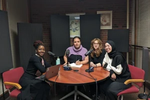 Four young people sit around a round wooden table. From left to right are a Black woman in a black dress, a Hispanic man in a purple top, a white woman with glasses and red hair, and a Middle Eastern woman wearing a black head scarf. All are smiling at the viewer.