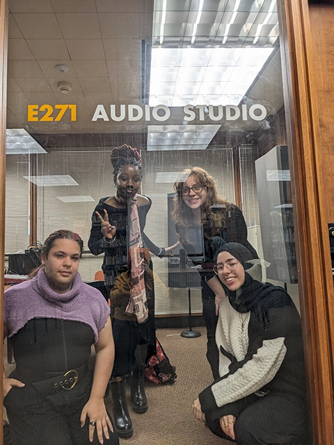 Four young people are posed behind a glass door with writing on it that reads "E271 Audio Studio" in gold and white letters. Sitting on the floor on the left is a Hispanic person in a purple crop top and black shirt. Standing behind them is a Black woman wearing a black outfit and colorful scarf giving a peace sign. Standing on the right is a white woman with black glasses and curly red hair. Kneeling on the floor on the right is a Middle Eastern woman wearing glasses, a white blouse, and a black head scarf.