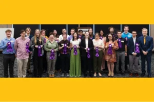 A large group of young people of varying races, all dressed in business casual, stand in a line and smile at the viewer. They each hold a purple show ribbon.