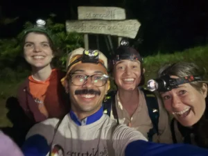 Four young people stand in front of a trail sign. It is dark and all four are wearing headlamps and are smiling at the viewer.