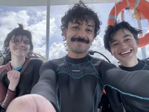 Three young people with damp hair and wearing wet suits smile in a boat. Behind them them is an orange life preserver.