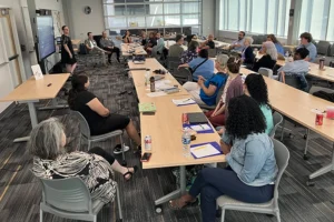 A large group of people sits around several long tables, all facing toward a presenter at the far end of the room.