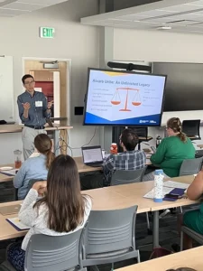 A man with black hair, glasses, and a button-down shirt stands at the front of a room next to a projector showing a PowerPoint slide. He talks to a group of people sitting at tables before him.