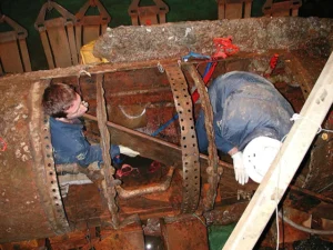 Two men sit inside a rusted metal tube with one side cut out to expose the interior.