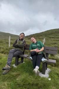 A middle-aged man and a younger white woman sit smiling on a brown bench. Rolling green hills and a gray, cloudy sky are in the background.