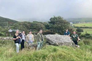 A group of six young people stand around a large rock in a grassy field. Behind them are rolling green hills and a blue-gray cloudy sky.