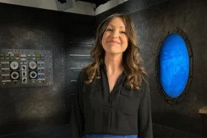 A white woman with long brown hair and a black shirt smiles at the viewer. Behind her are gray walls, one with a blue circular window and the other with a design of an instrument panel.