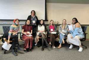 A group of six young women in business casual clothes sit in a line of a chairs and smile at the viewer. An older woman stands proudly behind them.