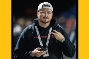 Headshot of a young man in a backward ball cap and sweatshirt with a press pass around his neck.