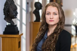 Headshot of a young woman with long curly brown hair. In the background are metal busts of heads on pillars.