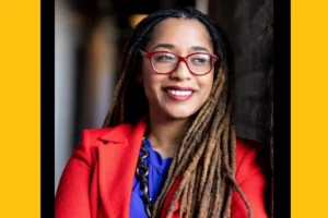A headshot of a young black woman with long hair, wearing red-framed glasses and a coral blazer.