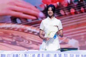 A young woman wearing white and holding a Chinese fan stands in front of a colorful background.