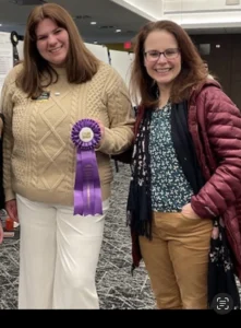 Two women, a student and teacher, stand together smiling. The student holds a purple award ribbon.