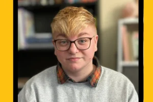 A headshot of a young person with short bleached hair and glasses