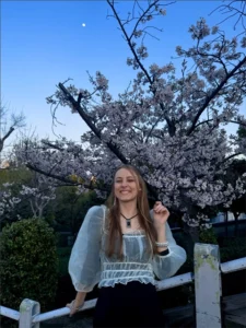 A young woman with long blonde hair wearing a white blouse stands in front of a cherry tree in full bloom.