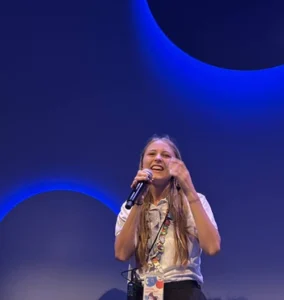 A young white woman with long blond hair and a white polo shirt holds a microphone and gestures at an unseen audience.