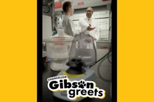 A young white man and an older Black man, both wearing white lab coats, converse in a laboratory. Along the bottom of the image are the word Chancellor Gibson Greets.