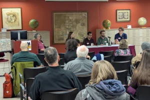 Four people sit behind a table at the front of a room. In the foreground in front of the table a crowd of people sits with their backs to the viewer, looking at the speakers.
