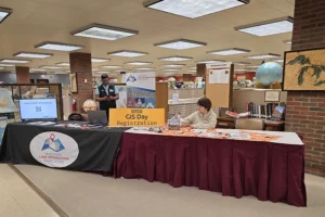 Two people stand behind a table that bears a sign reading GIS Day registration.