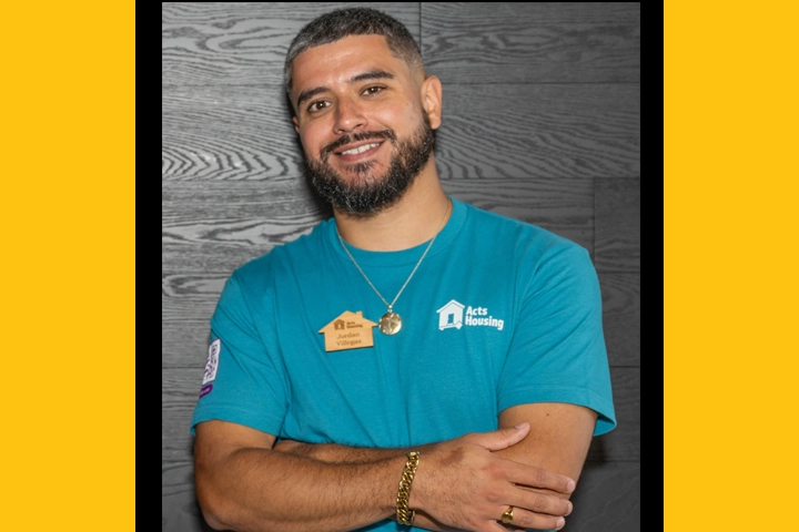A headshot of a Hispanic man with short dark hair wearing a teal t-shirt with an Acts Housing logo.