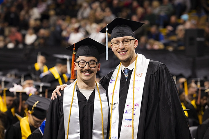 Two young men in black graduation robes with white stoles and gold cords pose and smile together in a crowded auditorium.