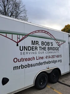 A white trailer is emblazoned with a bridge and the words Mr. Bob's Under the Bridge Serving Our Community, Outreach Line: 414-207-6615, mrbobsunderthebridge.org