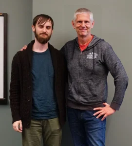 A middle-aged man has his arms around the shoulders of a younger man as the two smile at the viewer.