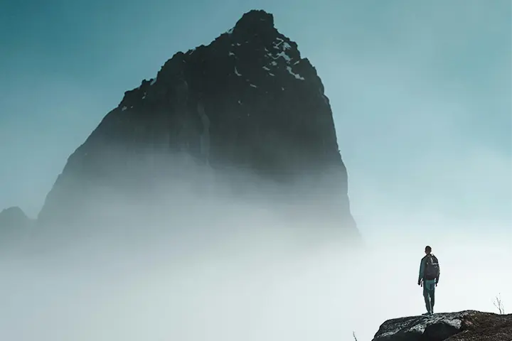 a person in shadow stands on a rock and looks up at a mountain surrounded by fog
